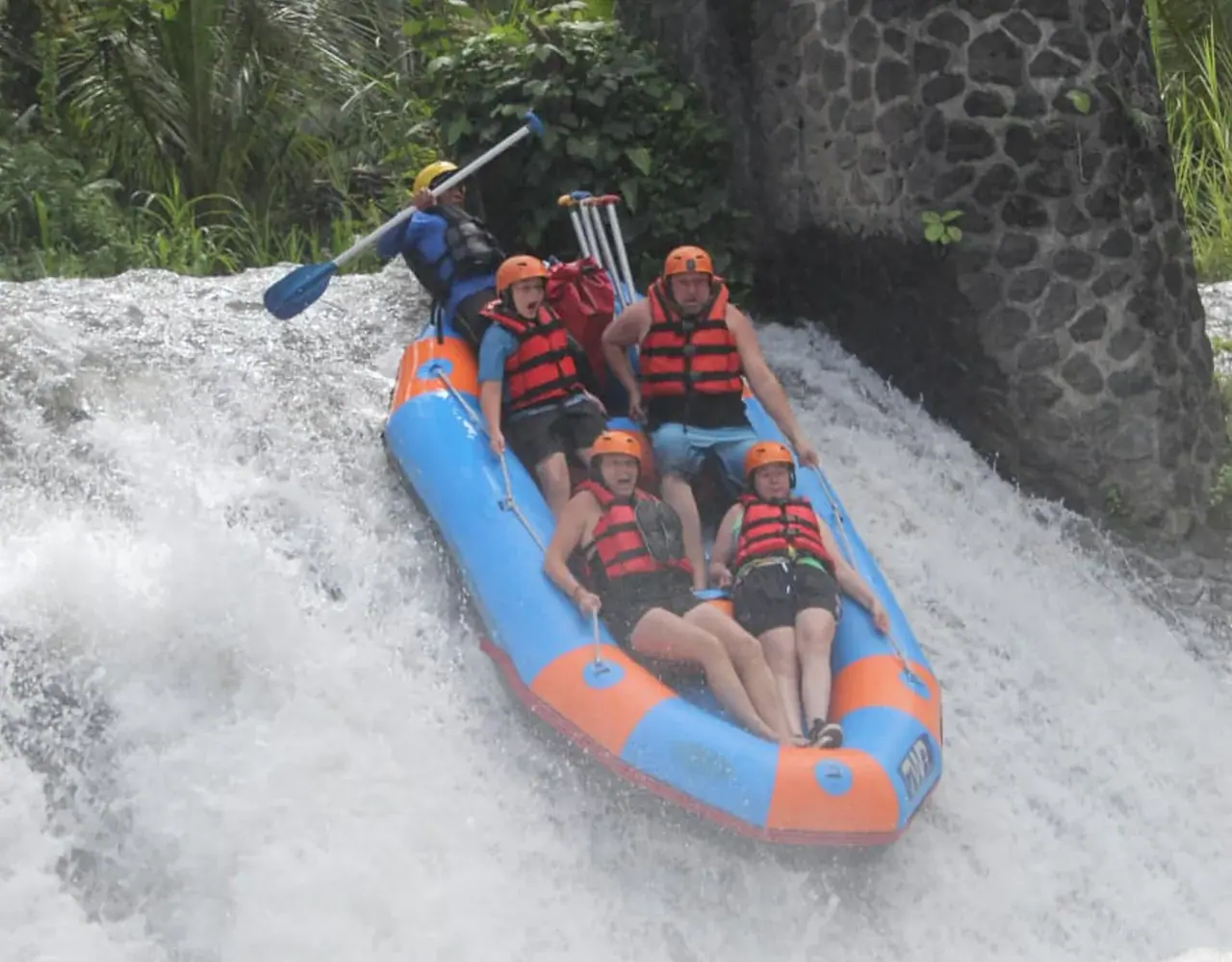 A blue and orange raft with four guests tipping over the edge of the dam drop on the Telaga Waja River
